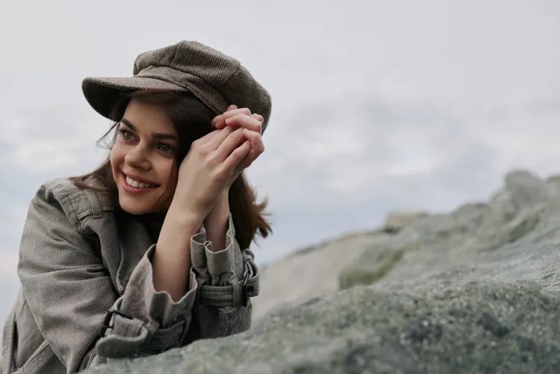 Smiling young woman wearing a patterned coat and matching cap leans on a rocky surface outdoors. The cloudy sky background creates a natural and relaxed mood in the image.