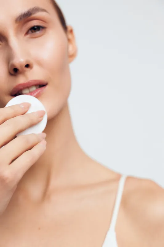 Woman skincare closeup, cotton pad on face, gentle beauty care routine, spa hygiene, moisturized skin texture, soft light, calm expression, minimal background