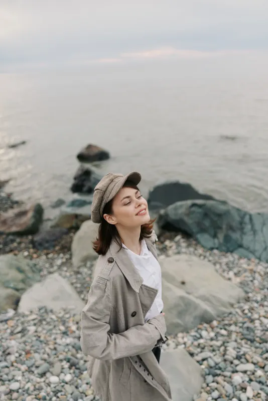 Young woman in beige coat and hat enjoys fresh air with closed eyes on rocky beach near calm sea. Concept of relaxation, peace, nature, and outdoor lifestyle at coast.