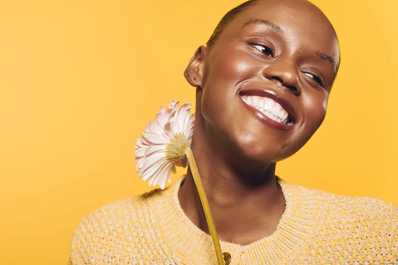 Smiling black woman with dark skin holds a pink flower near her neck against a yellow background. Concept of beauty, happiness, natural skincare, and positive mood with floral elements.