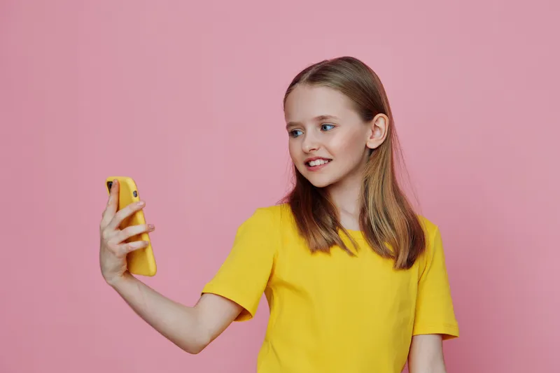 A cheerful young girl with light brown hair taking a selfie on a yellow phone against a pink background, portraying happiness and confidence