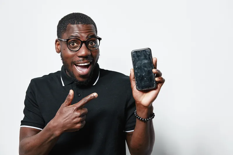 African American man with dark skin and glasses smiling and pointing at smartphone screen in black polo shirt on white background. Excited male showing mobile phone technology.
