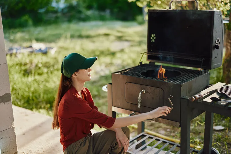 Young woman in casual clothes enjoying outdoor grilling, wearing a green cap, surrounded by greenery, grilling healthy food in a sunny environment