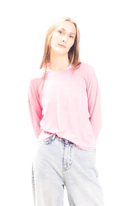 Young woman with long hair wearing pink sweater and light denim jeans stands against white background. Casual fashion portrait with soft natural lighting and relaxed mood.