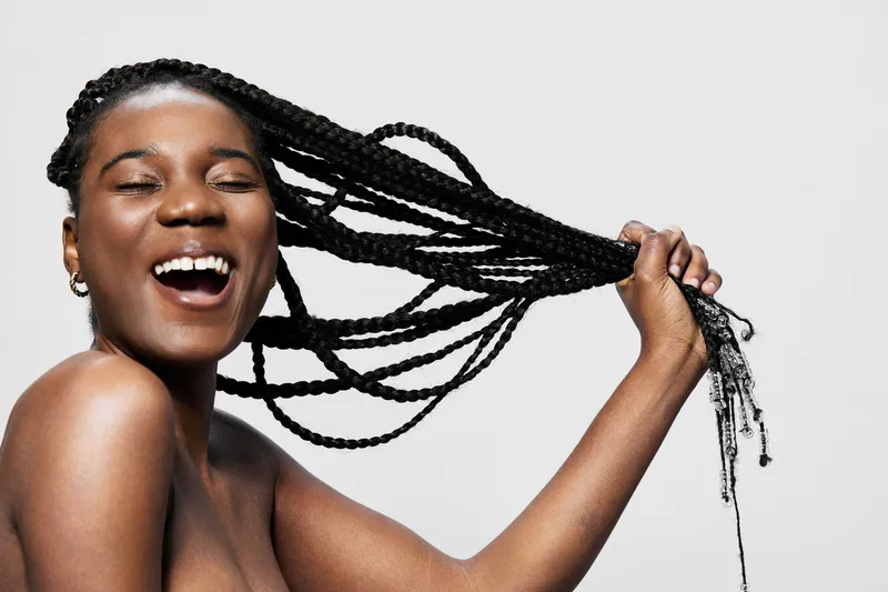 Happy African woman with long braids joyfully holds her braided hair, showcasing confidence and beauty in a bright and minimalist setting