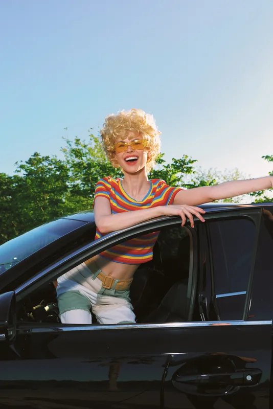 A joyful woman leans from a glossy car window, wearing a striped top and sunglasses, under bright sun. A candid lifestyle moment with retro color and carefree mood.