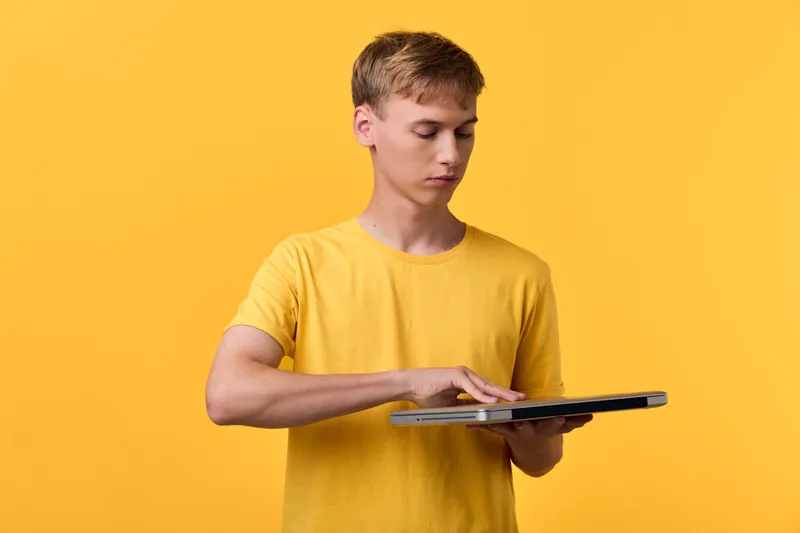 Young man in a yellow shirt taps a tablet on a touchscreen, focused and calm, against a bright yellow backdrop, illustrating modern device use and digital learning in a studio setting.