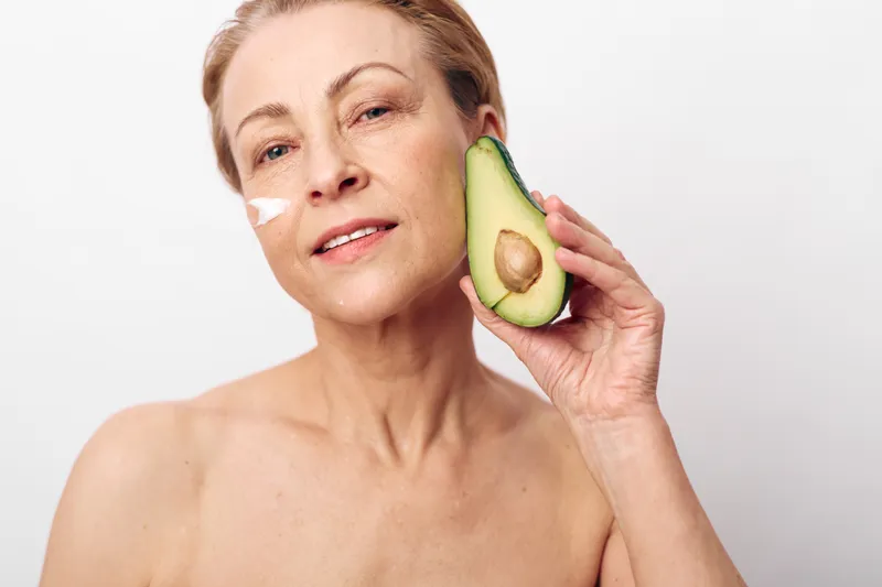 Mature woman with natural skin care cream on face holds half avocado near cheek. Concept of healthy aging, organic skincare, beauty routine, and natural facial treatment.