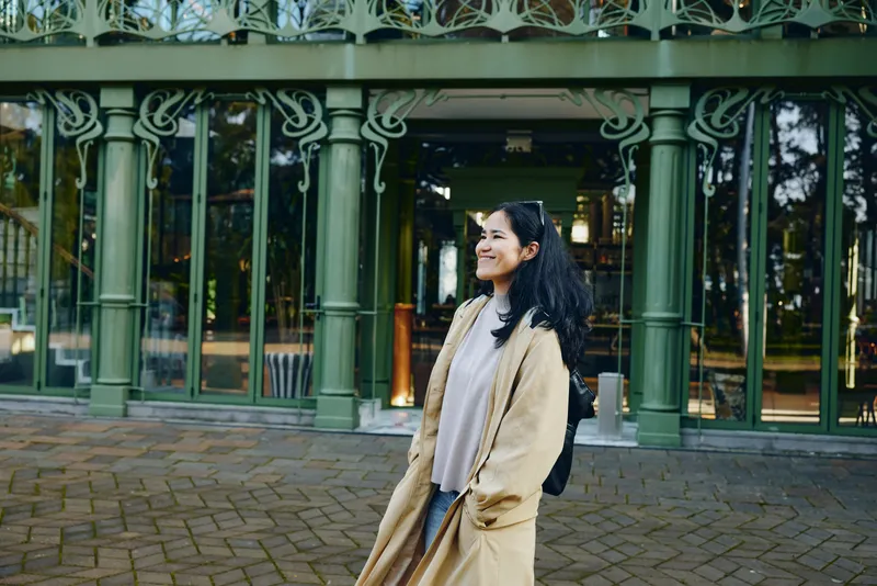 Woman in a long beige coat smiles outdoors in front of an ornate green storefront, casual fashion, urban street scene, lifestyle photography highlighting confidence and style