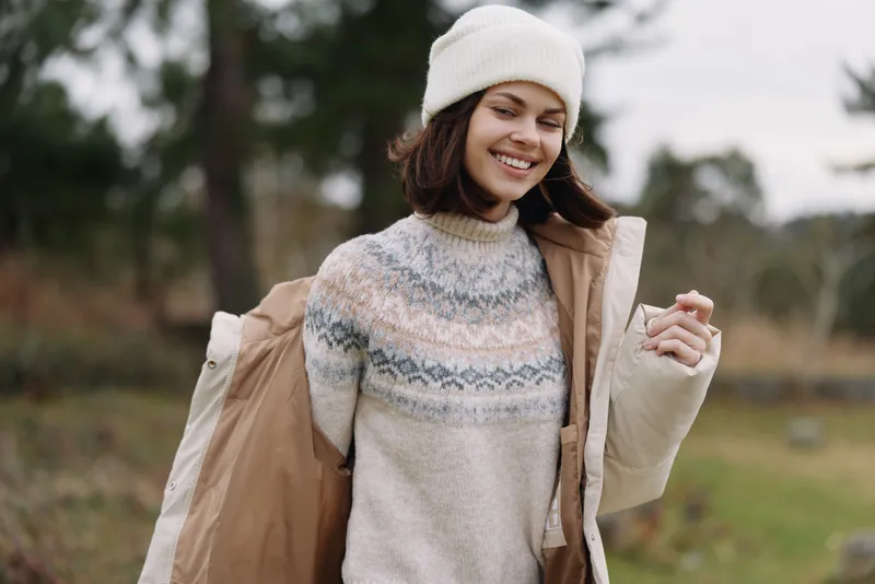 Woman smiling outdoors in a cozy knit sweater, white beanie and light jacket, carrying shopping bags in a park with blurred trees and a peaceful park.