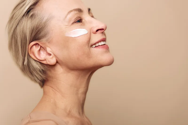 Mature woman with short blonde hair applies white face cream on cheek smiling gently in soft studio light. Concept of skincare routine, aging skin care, beauty and wellness.