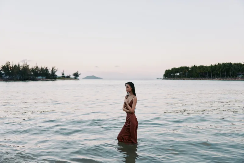 A woman stands waist-deep in calm sea water at sunset, wearing a flowing red dress that creates a graceful silhouette against the soft horizon and reflective water.