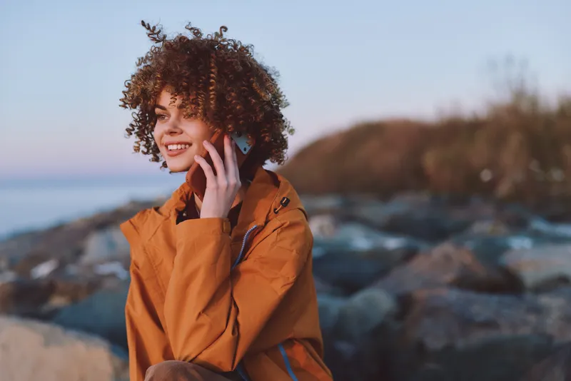 Woman lifestyle outdoors by the coast, curly hair, smiling while holding a phone to her ear, casual jacket, warm twilight light, relaxed conversation, shallow depth of field