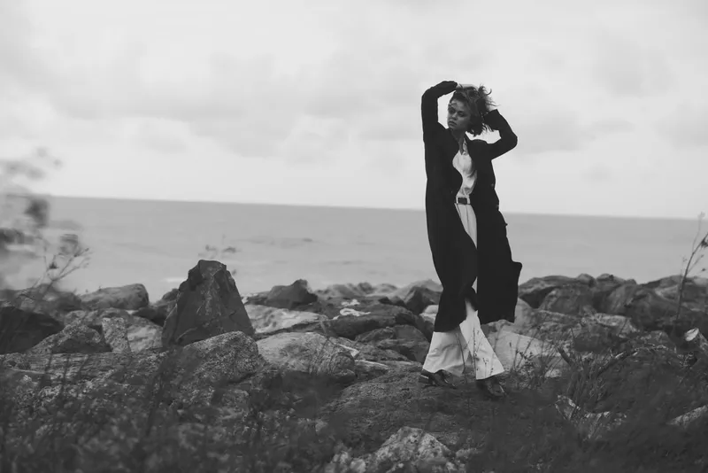 Woman on rocky coast in black and white, draped coat and flowing dress, windswept pose capturing dramatic fashion mood against rugged shoreline and sea horizon