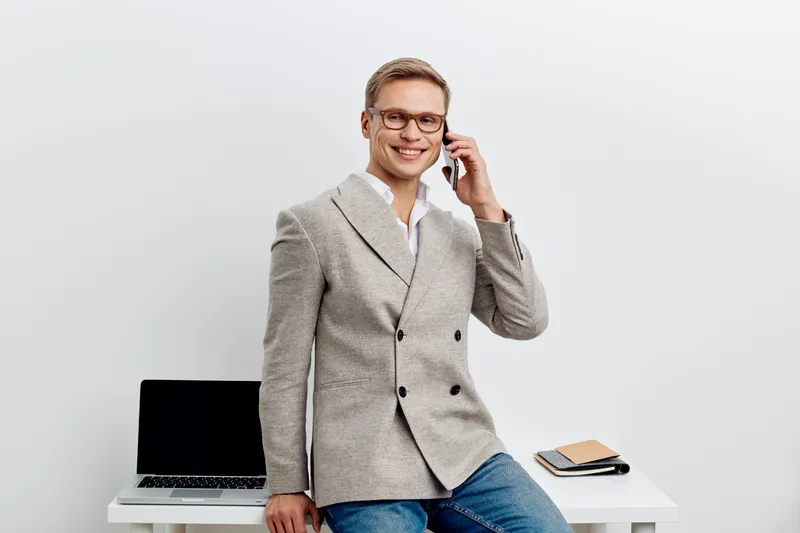 smiling young man talking smartphone wear casual blazer glasses sit near laptop desk isolated on white background work in digital nomad lifestyle concept
