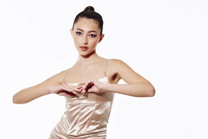 Young elegant woman with flawless makeup posing in shiny satin slip dress against a white backdrop. Her gaze is direct, her expression bold. Beauty fashion contemporary style concept.