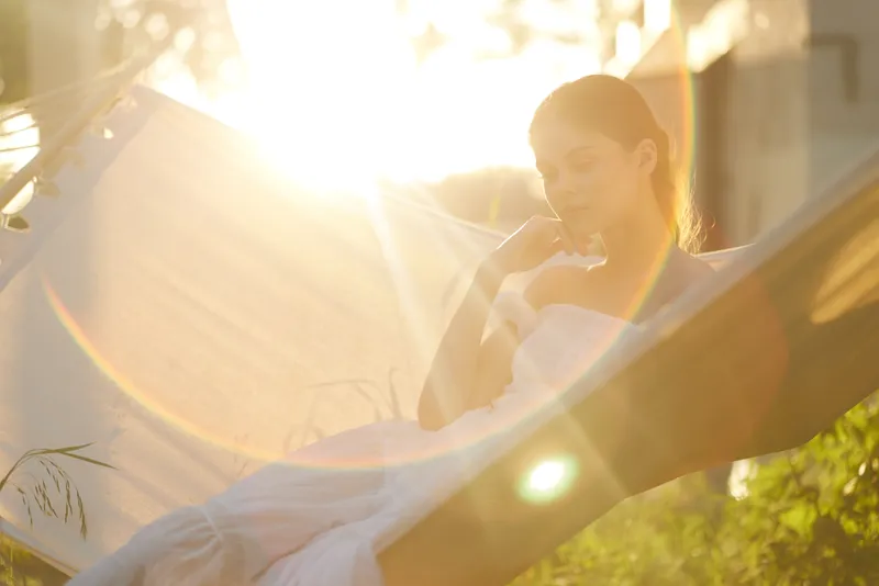 Young woman in a white dress relaxing in a hammock under sunlight, embodying tranquility and serenity in nature, creating a warm summer atmosphere