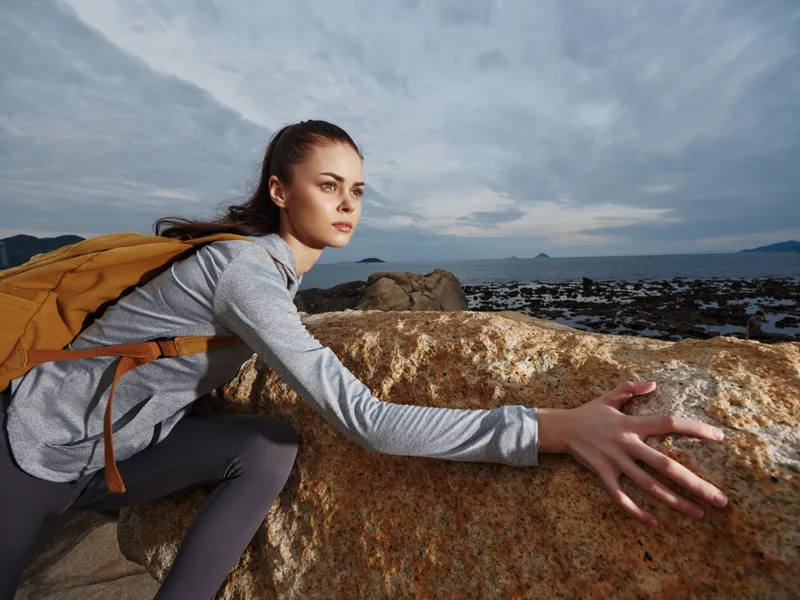 Adventurous young woman climbing rocks near the ocean, showcasing determination and outdoor spirit with a stylish backpack and scenic cloudy background