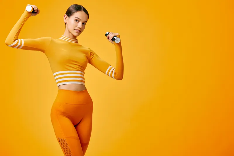 Female fitness athlete lifting small dumbbells in orange monochrome studio, wearing matching crop top and leggings; dynamic sport pose showing strength, form and energetic wellness.