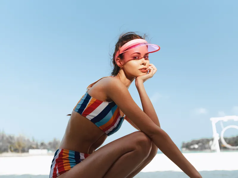 Young woman sits on a sunny beach wearing a striped bikini and pink visor, gazing thoughtfully with her chin resting on her hand, radiating relaxed seaside charm.