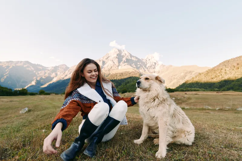 Woman and dog in mountains meadow, petting companion and smiling during outdoor nature excursion. Grassy field, scenic peaks, relaxed happy portrait of friendship and freedom.