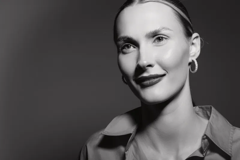 Portrait of a smiling Caucasian woman with light skin and slicked-back hair wearing a shirt and hoop earrings. Studio lighting highlights her clear complexion and confident expression.