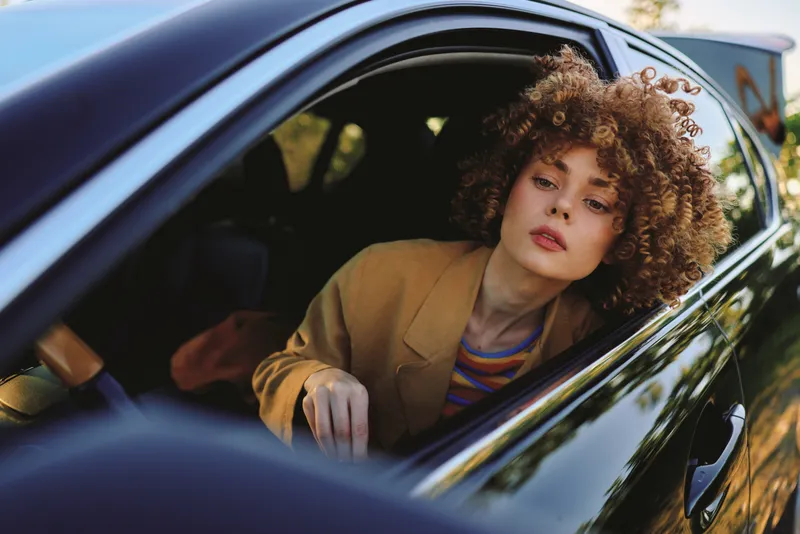 Curly haired woman leaning from a car window, casual outfit, moody daylight. Scene evokes lifestyle photography, carefree travel, vintage vibe, and bold personality, with soft shadows and warm tones.