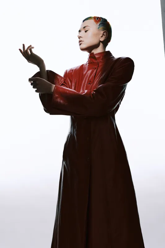 Woman in red leather coat poses gracefully against bright white background, showcasing stylish modern fashion with artistic colorful hair detail, minimalist studio setting