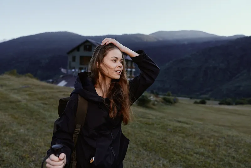 A young woman hiking outdoors in a rugged landscape, wearing a jacket and backpack. She smiles softly, lifts a hand to her hair, and enjoys the fresh air and expansive, open scenery around her.