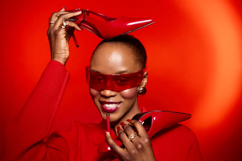 Elegant black woman with dark skin wearing red sunglasses and red outfit holds red high heels and lipstick against bright red background. Concept of fashion, beauty, and style.