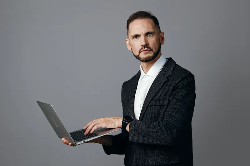 A poised business professional man stands with a laptop, presenting a focused and confident emotion against a clean isolated colored background.