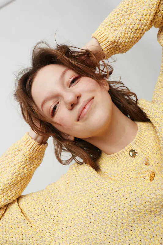 Smiling young woman with curly hair wearing yellow knitted sweater looks relaxed and happy. Close-up portrait on light background with natural expression and casual style.