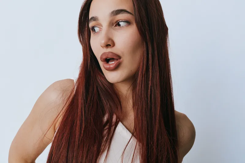 Woman with long red hair and expressive face looking to side wearing white top against plain background. Portrait of young female with natural makeup and casual style in studio.
