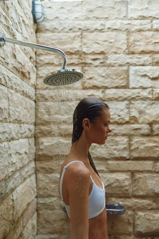 young woman in white top standing under outdoor shower with stone wall background, wet hair, side profile, natural light, summer freshness essence