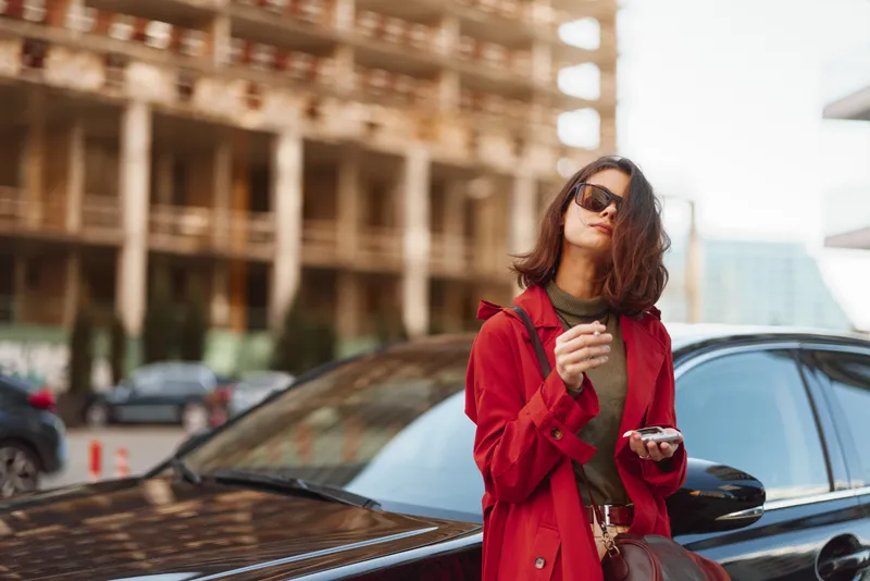 Young woman in a red coat stands confidently near a black car while holding a smartphone and wearing sunglasses in an urban setting.