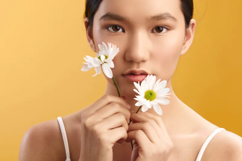 asian woman holds white daisies near her face against a warm yellow background, creating a natural portrait that conveys calm elegance and subtle celebration of simple beauty.