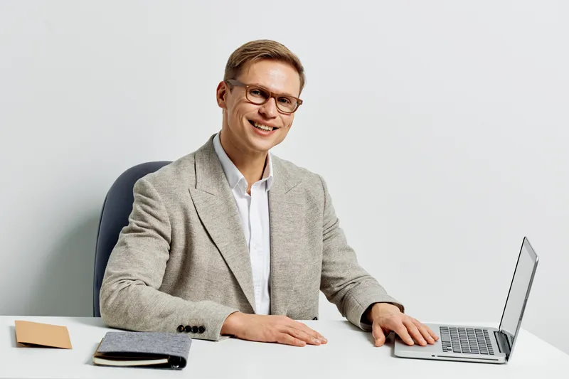 Professional smiling young Caucasian man wearing glasses in formal attire sitting at desk with laptop, notebooks, and pen isolated on plain white background studio portrait. Business professional conc