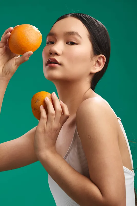 asian woman holds two oranges against a vibrant green backdrop, a fresh studio portrait that conveys vitality and calm confidence, with soft light enhancing skin tone and natural allure.