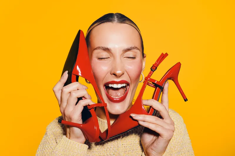Happy woman with closed eyes holds red high heel shoes near face on bright yellow background. Concept of fashion, style, footwear, and joyful emotions in studio lighting.