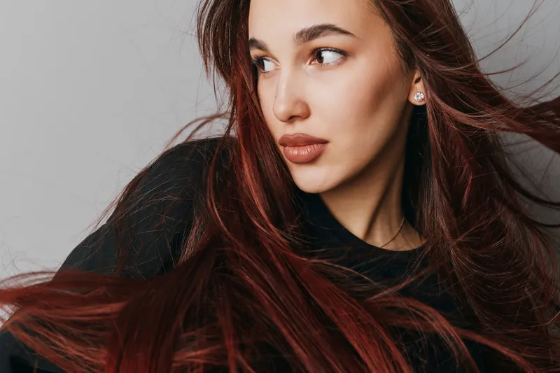 Young woman with long brown hair looking sideways in studio. Portrait of attractive female model with natural makeup and earrings on grey background with flowing hair movement.
