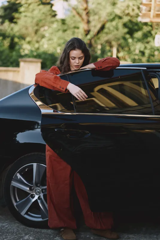 A woman in a red outfit leans on a sleek black car, her head resting on her arm as she gazes downward. A quiet, urban lifestyle moment highlighting contemplation and style