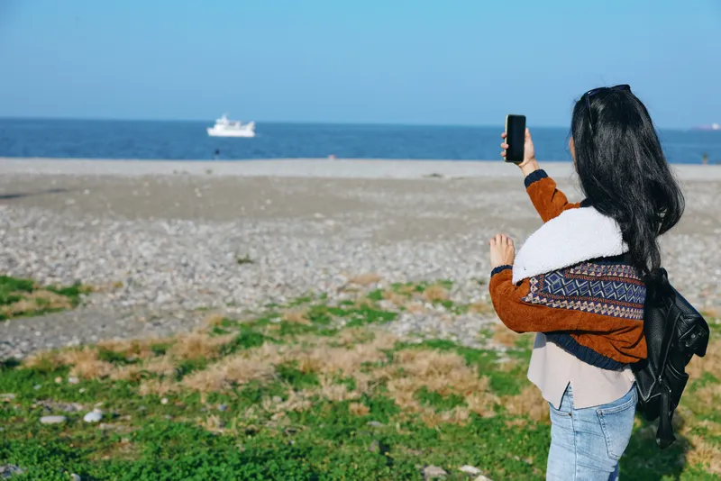 Woman with backpack captures seaside landscape on a sunny day, using a smartphone, standing on a grassy dune overlooking blue ocean and a distant boat