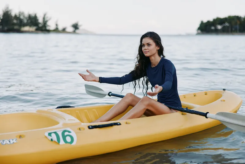 A young woman sits in a yellow kayak on calm water, paddles resting across the boat, gazing ahead with a focused yet relaxed expression under soft daylight.