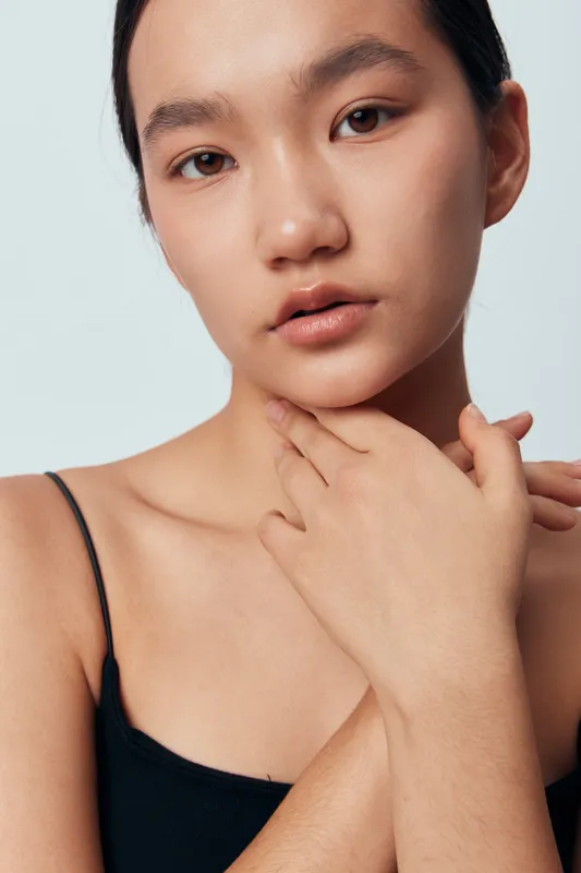Young woman with natural skin touching neck and shoulder, wearing black top in studio. Portrait with soft light, minimal makeup and calm expression for beauty and skincare concept.