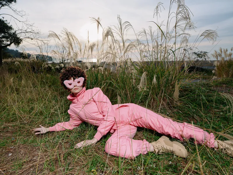 A person in a pink suit lies in tall grass outdoors, wearing a mask that resembles a doll. This surreal and eerie scene captures a strange, still moment amid reeds and a distant sky.
