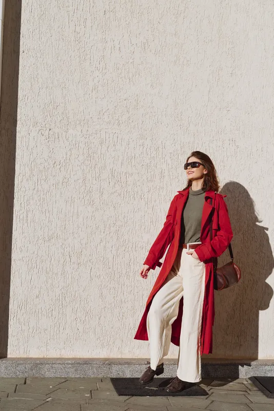 Stylish young woman in a red trench coat and white pants stands beside a textured wall, basking in daylight with a confident street style vibe.