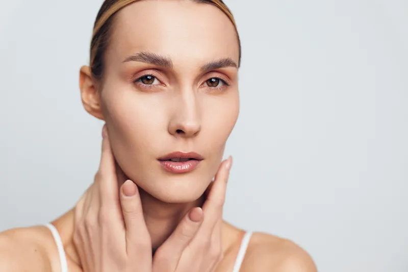 A close-up portrait of a young woman with clear skin gently touching her neck against a neutral background. Concept for skincare, beauty, and natural health care.