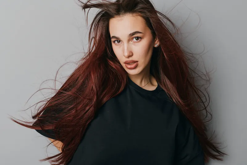 Young woman with long red hair and black shirt posing against gray background, looking at camera with confident expression and natural makeup in studio portrait.