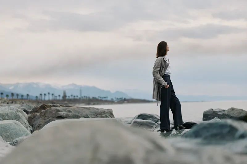 Young woman stands on rocks near a calm sea, looking into the distance. Mountains and cloudy sky create a peaceful outdoor scene with natural mood and solitude concept.