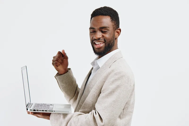 Smiling confident man holding a laptop with one hand, looking at camera and giving a thumbs-up gesture, wearing beige sweater and white shirt, isolated on plain white background, studio portrait, life